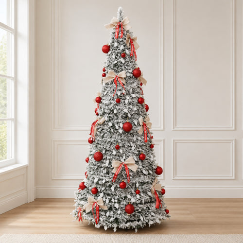 A tall, flocked Christmas tree decorated with red ornaments and beige ribbon bows, standing on a light wood floor against a cream paneled wall.