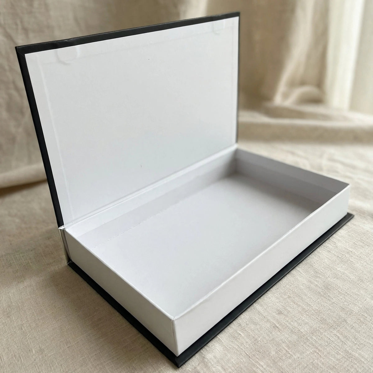 An open rectangular gift box with a white interior and black outer edges, photographed on a beige fabric surface with a softly blurred neutral background.