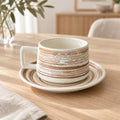 A white ceramic cup with a matching saucer, both decorated with a rustic brown distressed horizontal stripe pattern, set on a light wooden table in a cozy, softly blurred interior setting.