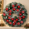 A festive Christmas wreath with green artificial pine branches, white frosted tips, red ornaments, striped pinecones, and small white berries, displayed on a mantel against a warm beige wall with candlelight in the background.