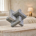 A gray concrete knot sculpture, resembling a modern wooden or stone jenga block, displayed on a light beige stone ledge in a softly blurred bedroom setting.