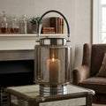 A ribbed glass lantern table lamp with a lit candle inside, featuring a dark gray rope handle and polished silver metal base, set on a mirrored coffee table in a cozy living room.