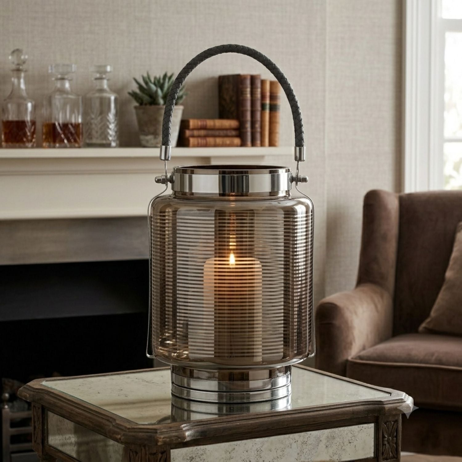 A ribbed glass lantern table lamp with a lit candle inside, featuring a dark gray rope handle and polished silver metal base, set on a mirrored coffee table in a cozy living room.