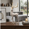 A white deer figurine with large gold antlers stands on a wooden table in a modern living room setting, with a gray sofa, framed art, books, and a candle in the background.