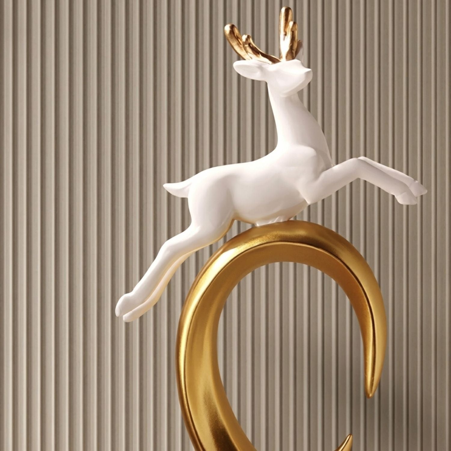 A decorative sculpture of a white deer with gold antlers perched on a curved gold base, set against a beige vertically ribbed background.