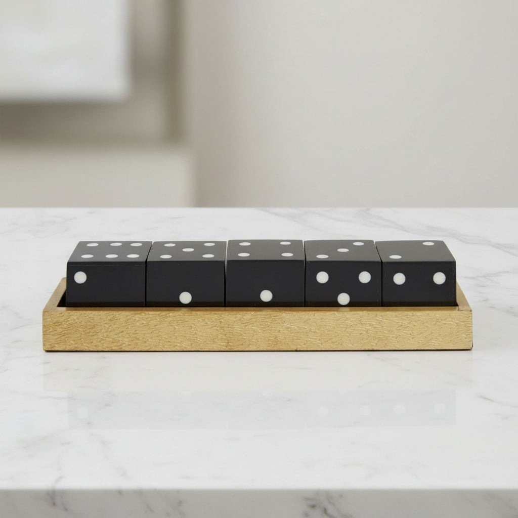 Five black dice with white pips resting on a rectangular light wood tray, placed on a white marble surface with a softly blurred neutral background.