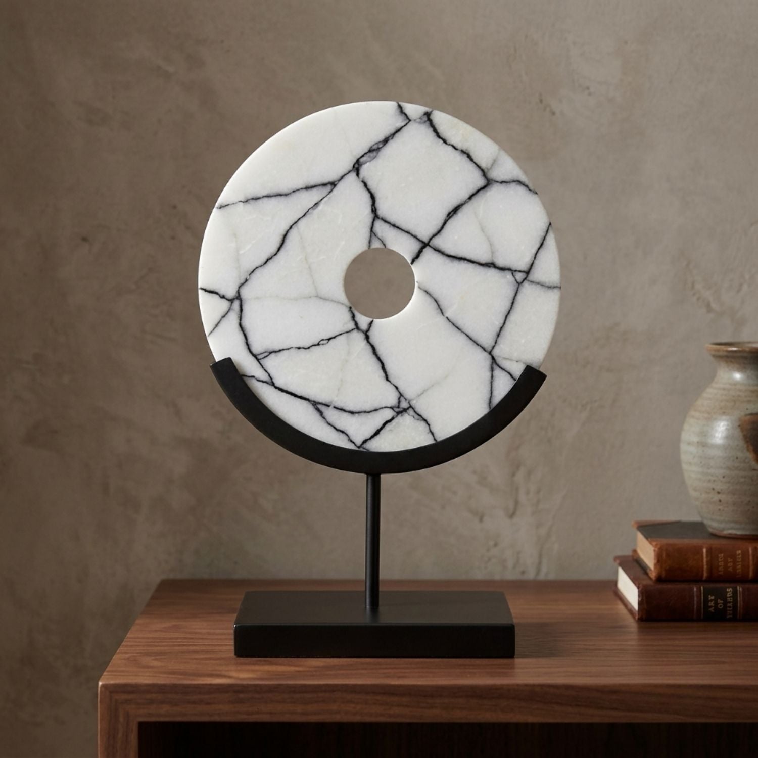 A round white marble tabletop sculpture with black veining, mounted on a black metal stand with a square base, displayed on a wooden table next to books and a ceramic vase.