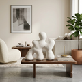 Three abstract white stone figurines displayed on a wooden coffee table in a modern, neutral-toned living room.