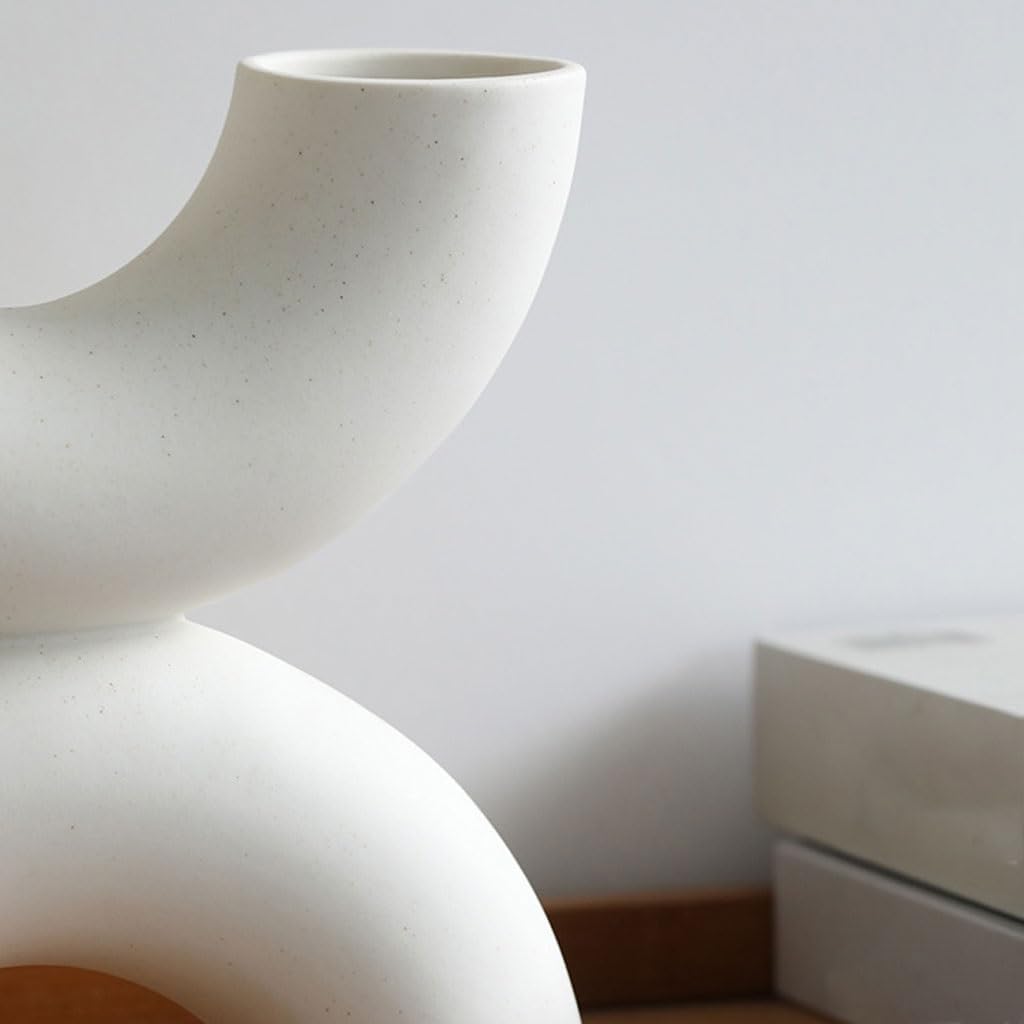 A close-up of a modern, sculptural ceramic vase in a matte off-white finish with subtle speckles, set against a minimalist light gray wall with a wooden base.