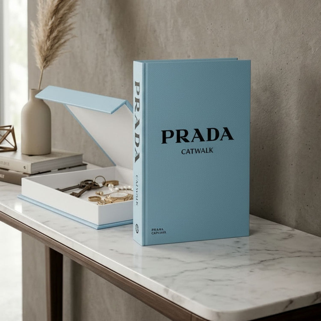 A light blue hardcover book or box set standing on a marble-topped table, with an open matching box beside it containing jewelry. The front cover reads PRADA CATWALK in black text, and the spine also shows PRADA.