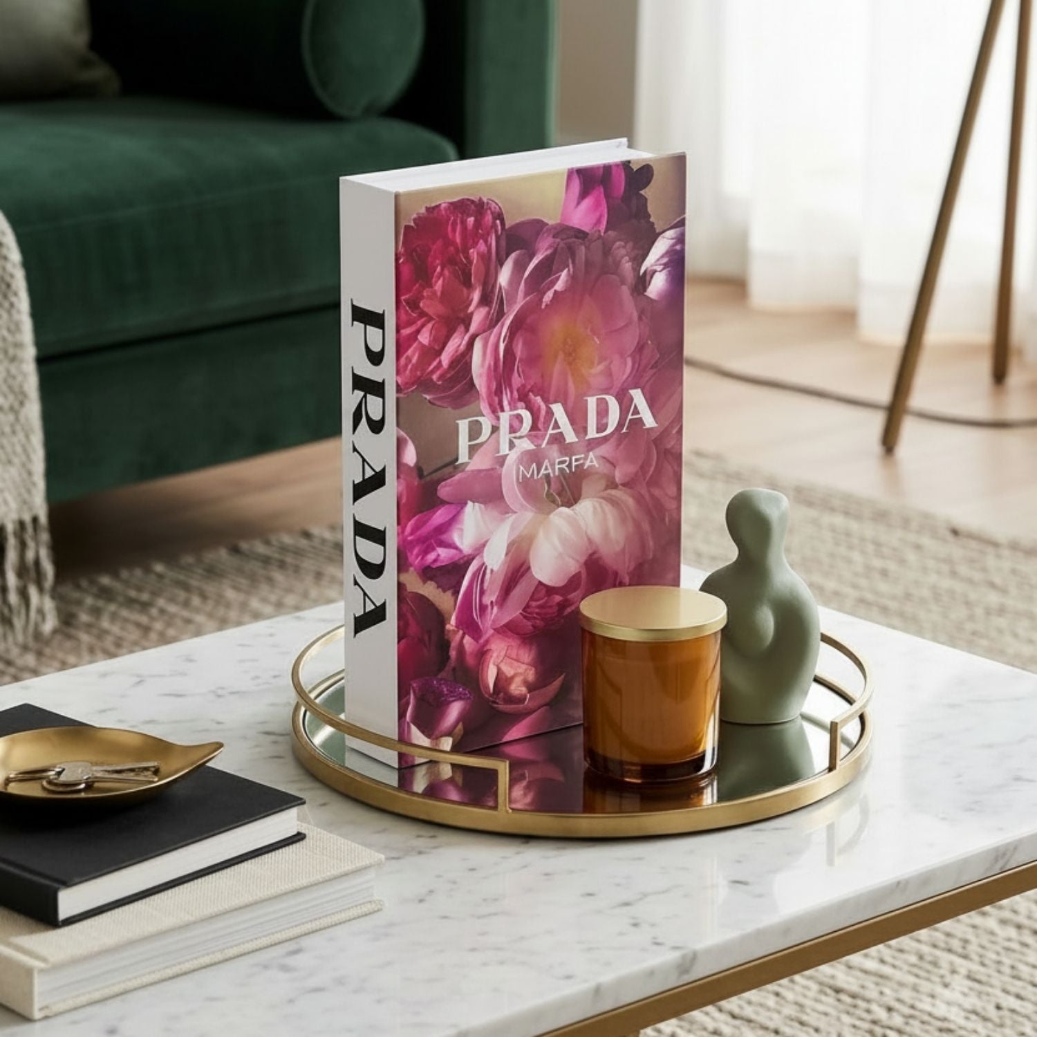 A hardcover book with a pink floral cover and vertical PRADA branding stands on a round gold-trimmed tray on a marble coffee table, styled in a modern living room setting.