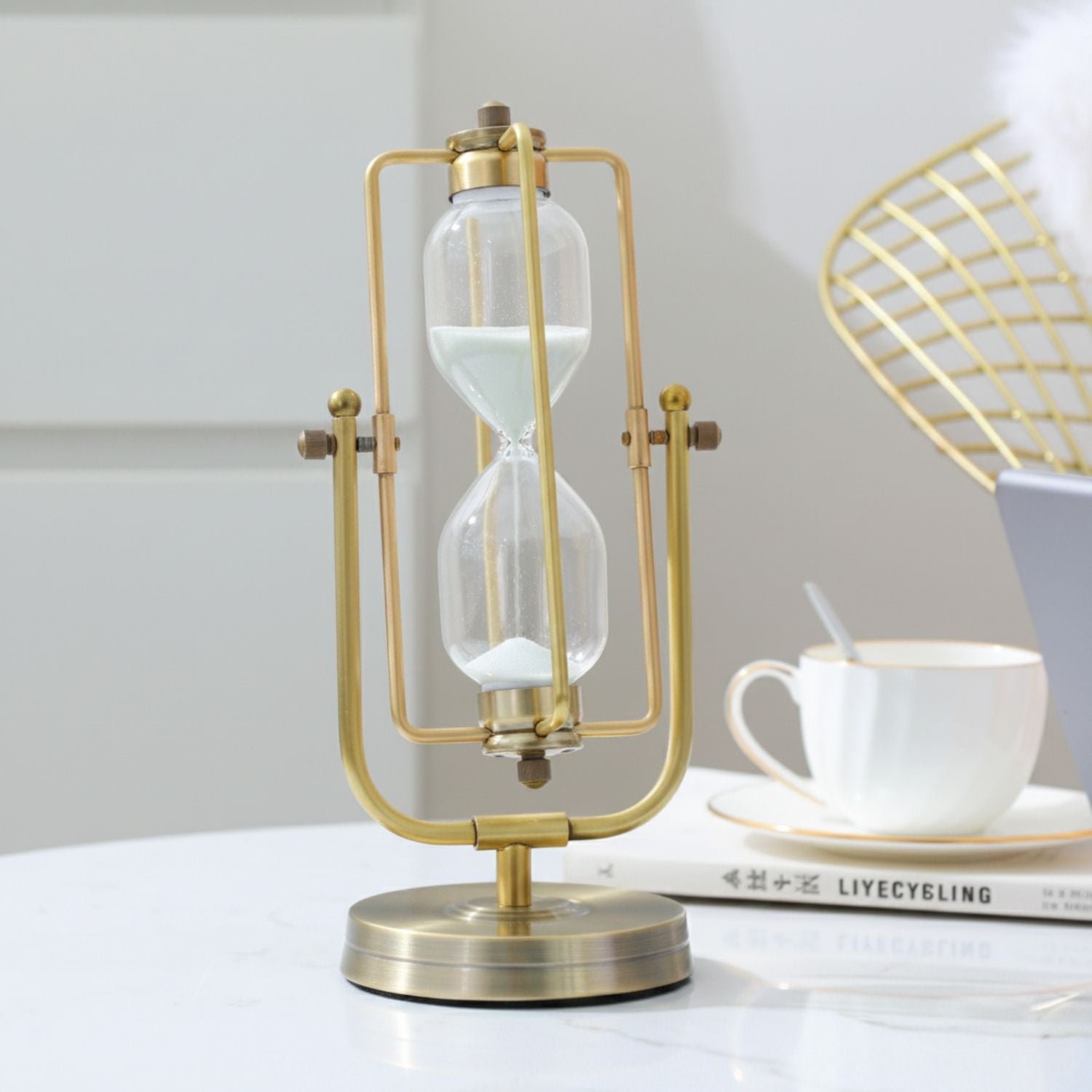 A gold-finished desktop hourglass with a clear glass bulb and white sand, styled on a white table next to a white teacup and a book.