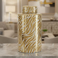 A tall cylindrical decorative canister with a matching lid, featuring a glossy gold and white zebra-stripe pattern, displayed on a marble countertop in a blurred living room setting.