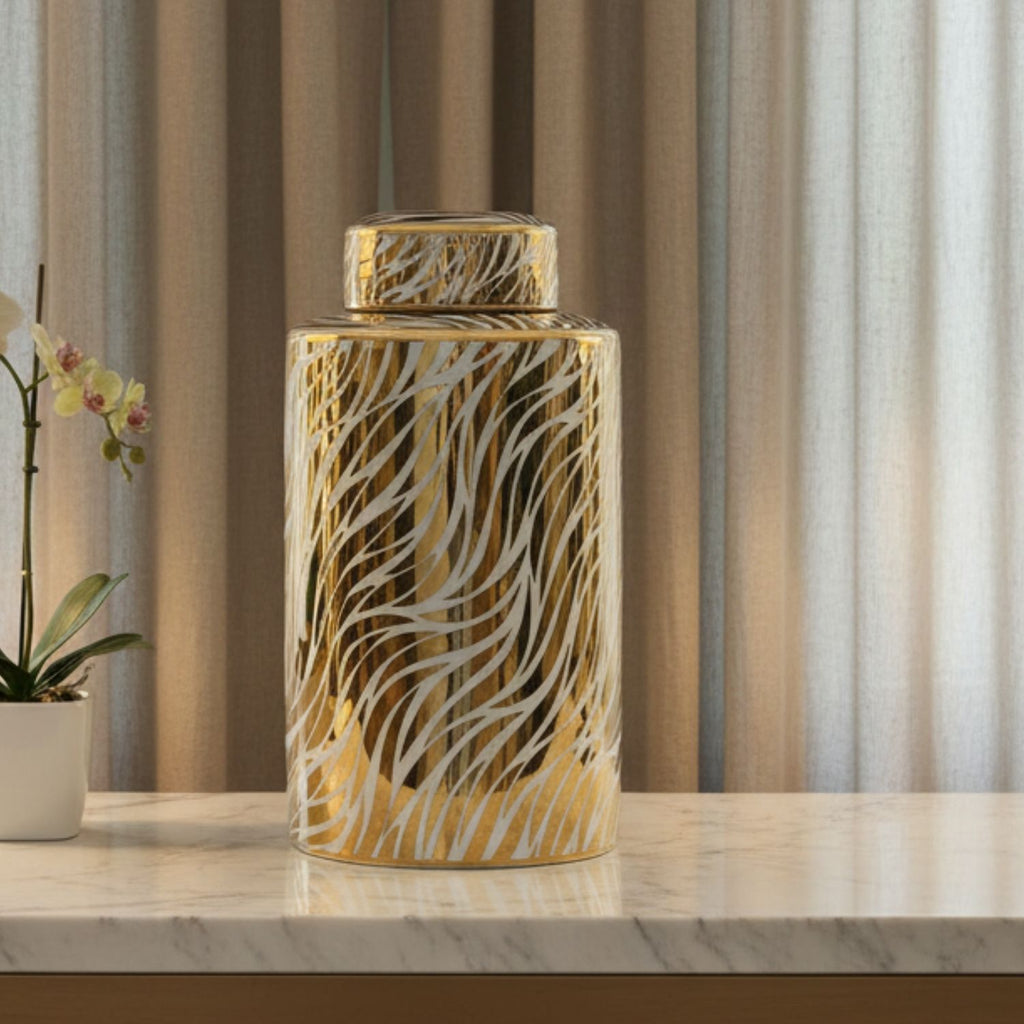 A tall cylindrical decorative jar with a lid, featuring a metallic gold and white wavy abstract pattern, displayed on a marble countertop in front of beige curtains.