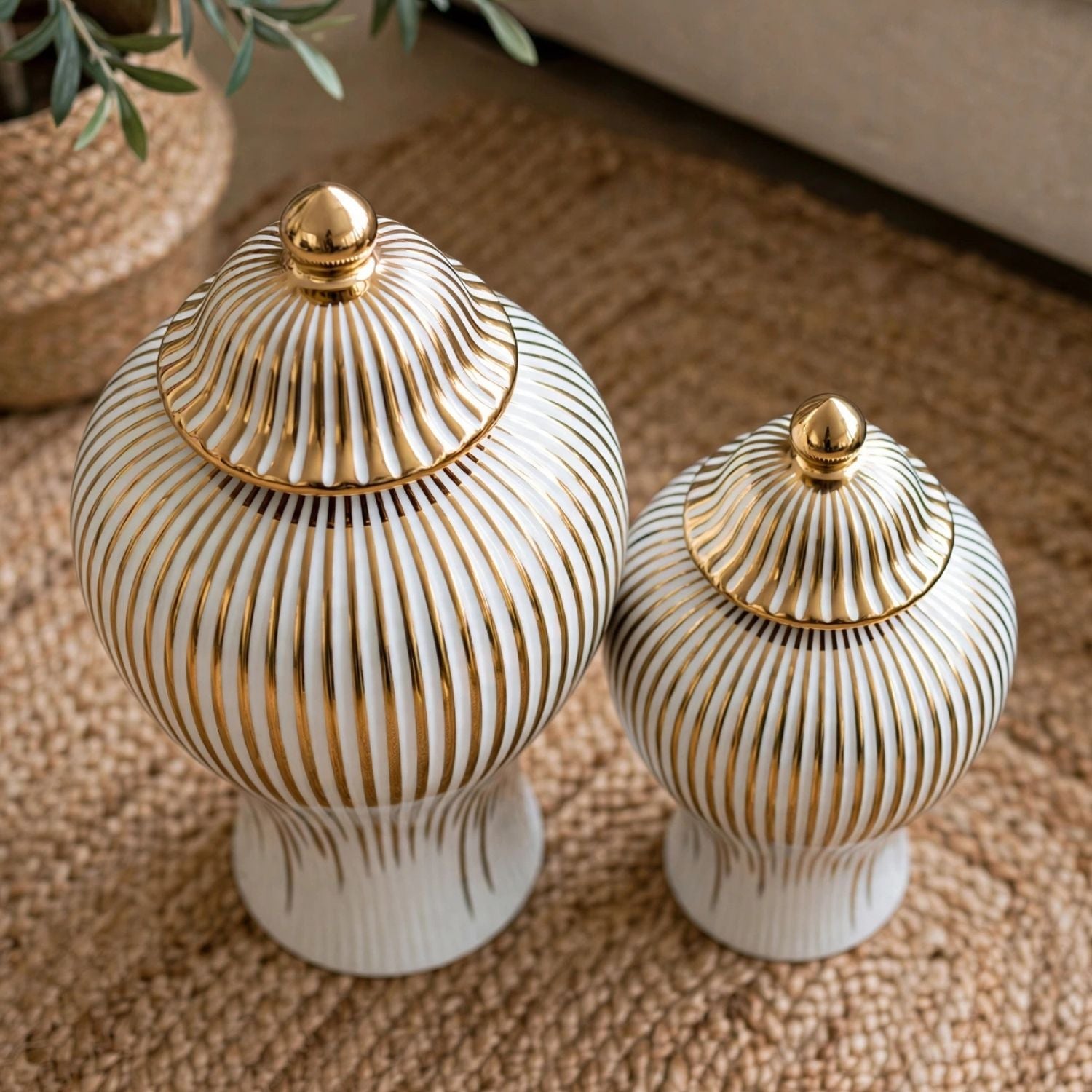 Two ribbed ceramic lidded jars in off-white with metallic gold vertical stripes and gold finials, displayed on a woven beige rug.
