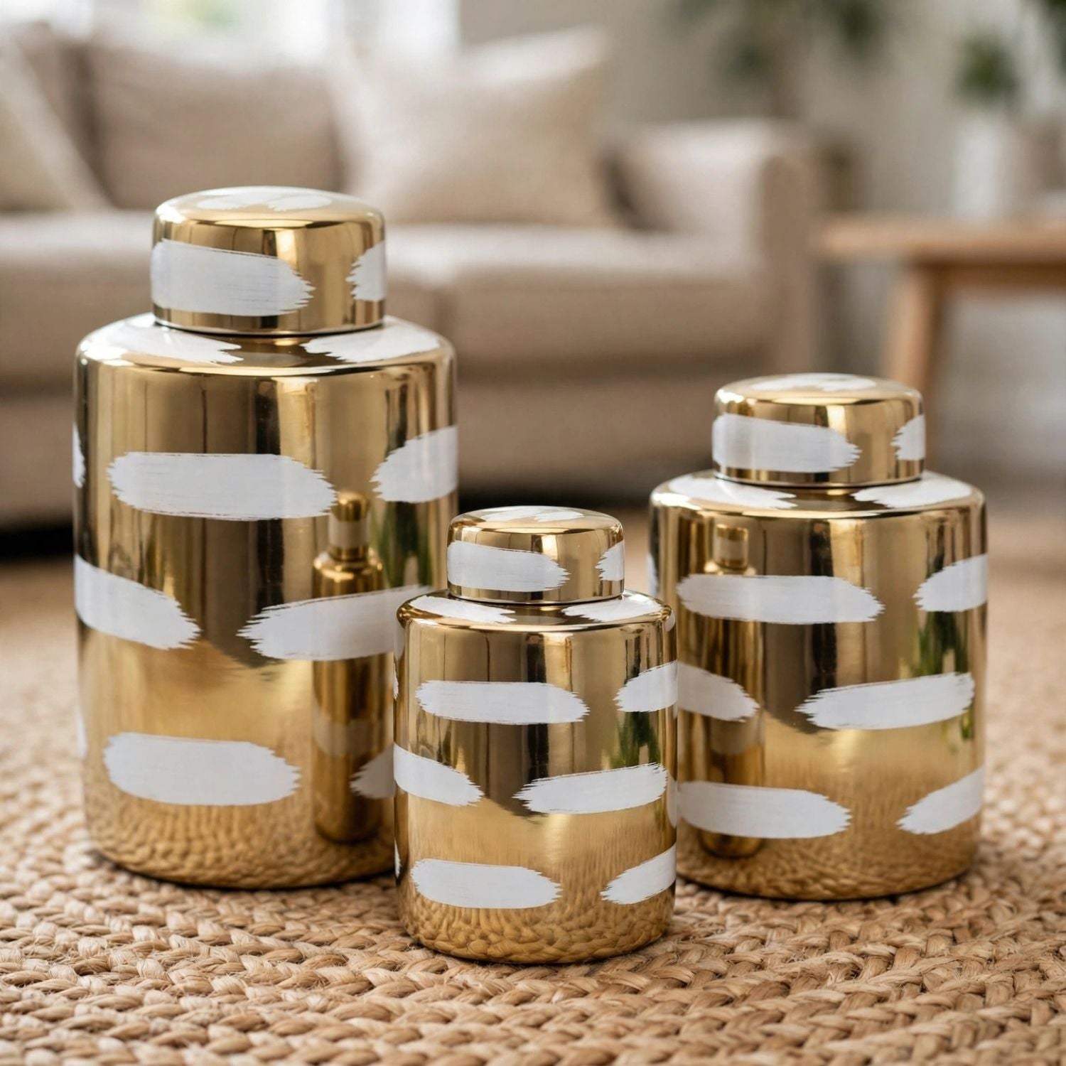 Three metallic gold decorative canisters with white brushstroke patterns, arranged on a woven beige rug in a softly blurred living room setting.