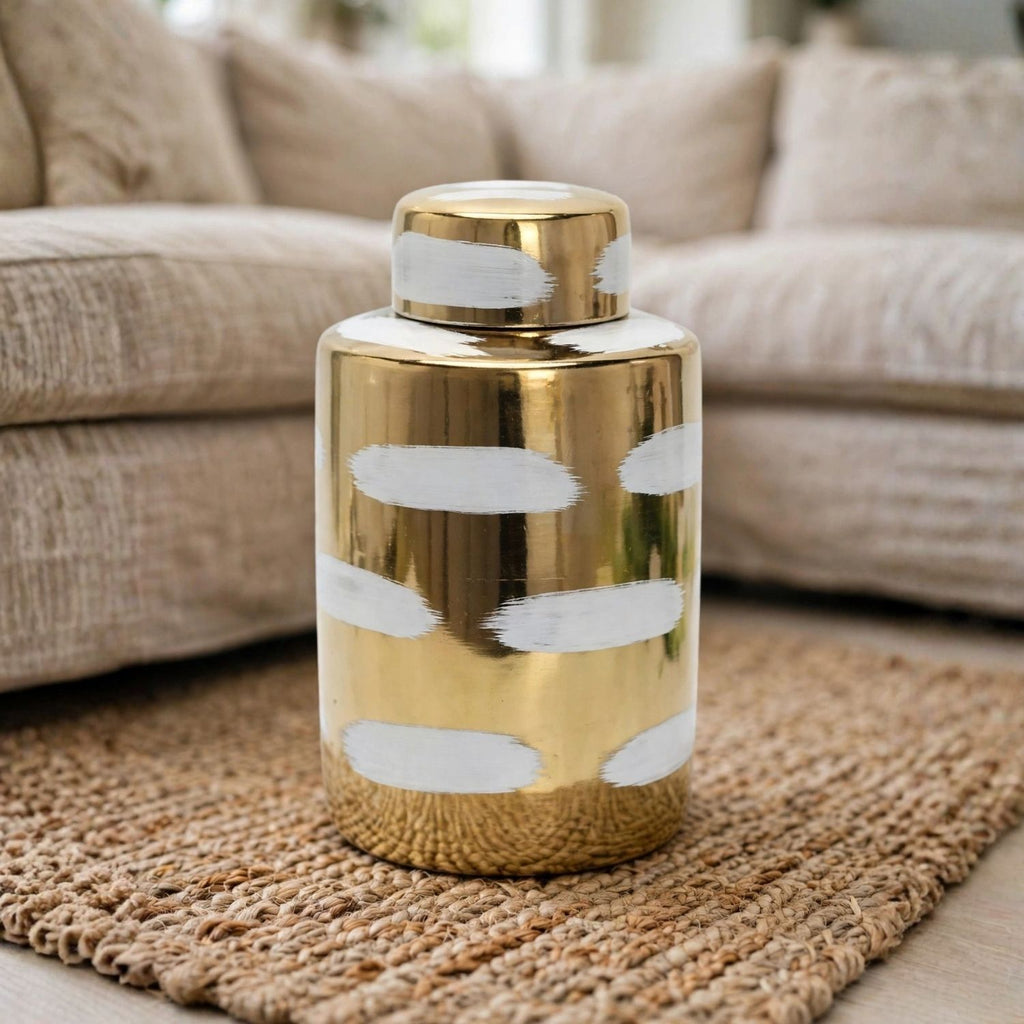 A cylindrical gold ceramic jar with a matching lid, decorated with white hand-painted brushstroke patterns, set on a woven jute rug in front of a beige sofa.