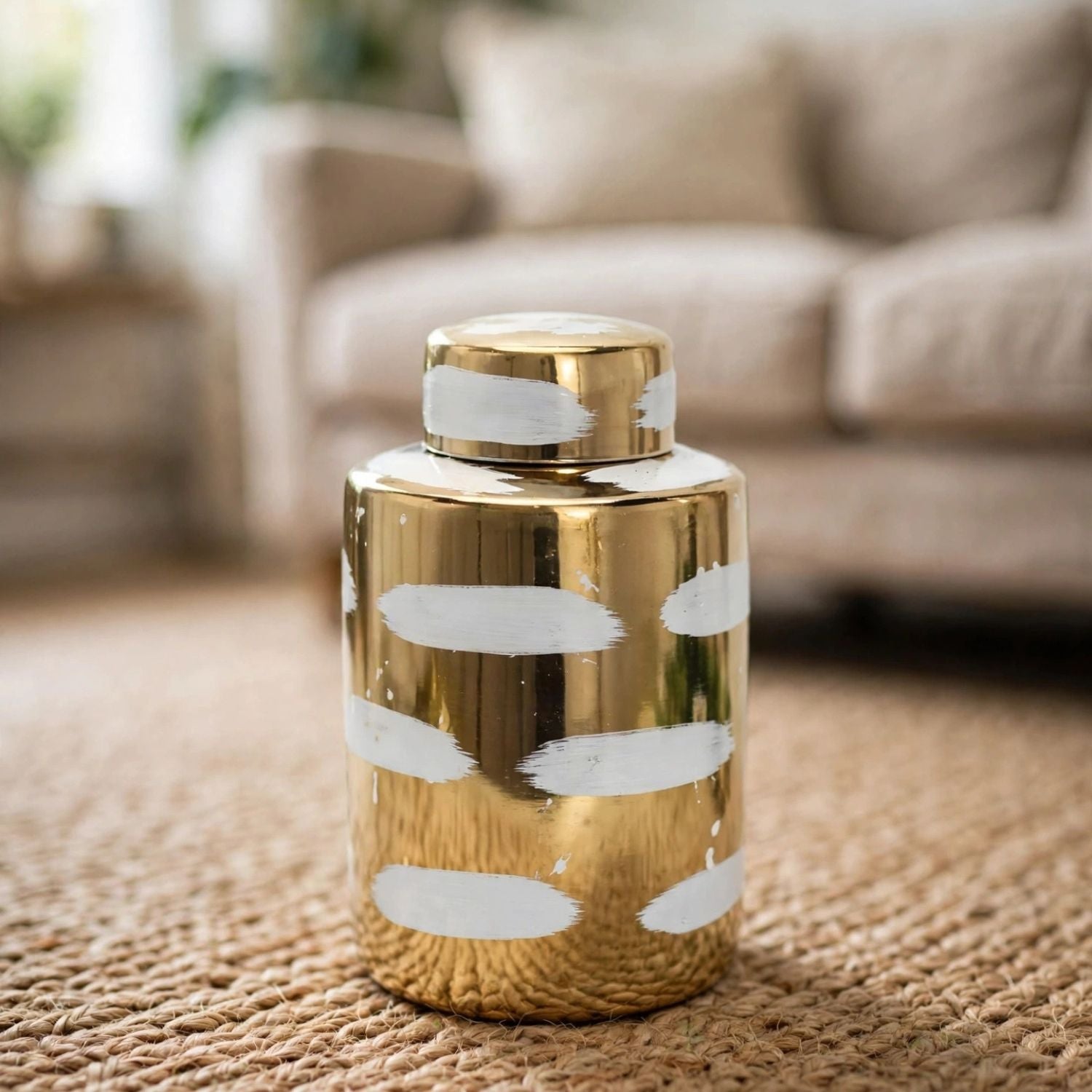 A shiny gold cylindrical decorative container with a matching lid, decorated with irregular white brushstroke patterns, set on a woven jute rug in a blurred living room background.