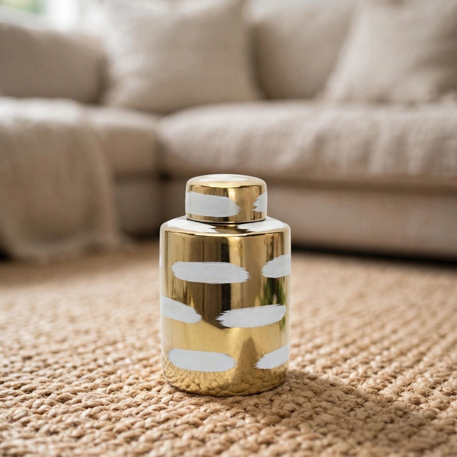 A small cylindrical decorative container with a shiny gold finish and white abstract brushstroke patterns, standing on a woven beige rug in front of a blurred sofa.