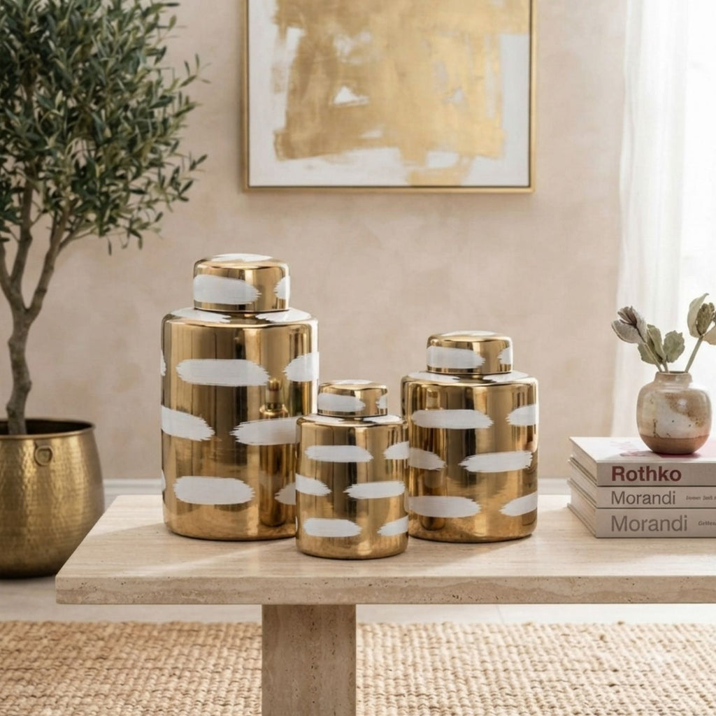Three metallic gold decorative canisters with white abstract brushstroke patterns, displayed on a light wood table in a styled interior setting.