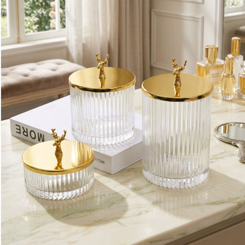 Three ribbed clear glass storage jars with shiny gold lids and small gold deer figurines on top, arranged on a marble surface in a bright room.