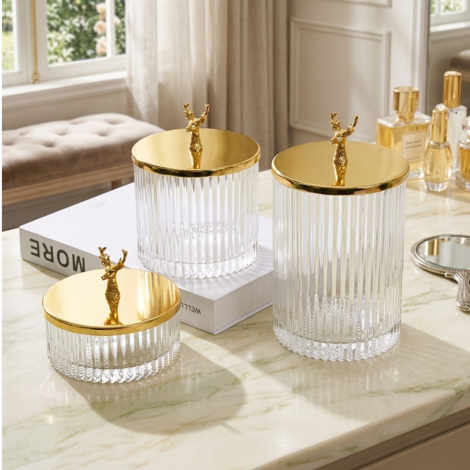 Three ribbed clear glass storage jars with shiny gold lids and small gold deer figurines on top, arranged on a marble surface in a bright room.
