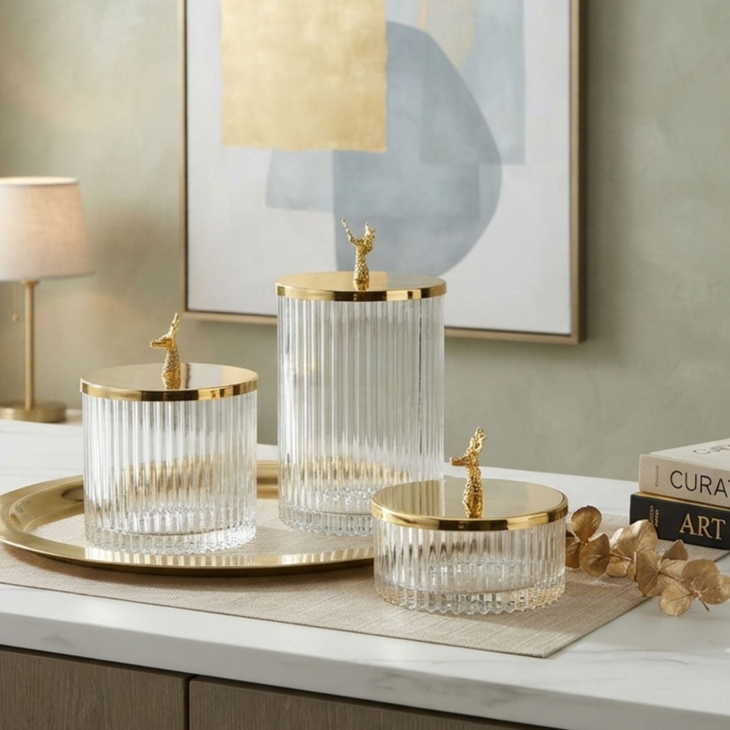 Three ribbed clear glass storage jars with gold lids and deer-shaped gold finials, arranged on a gold tray and beige runner on a white marble countertop in a styled interior setting.