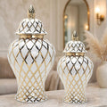 Two white ceramic lidded storage vases with gold and black diamond lattice patterns, displayed on a marble surface in a softly lit interior setting.