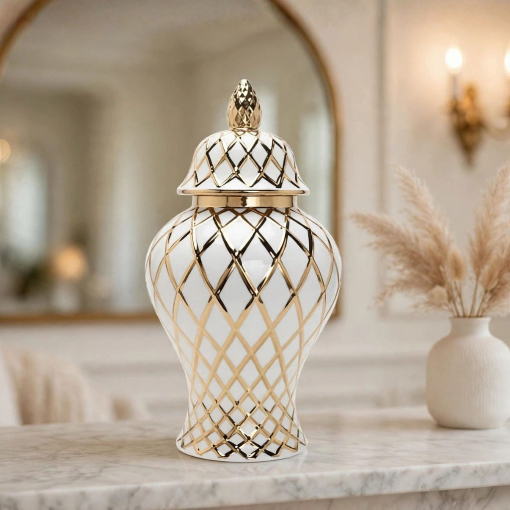 A white ceramic lidded jar with a gold geometric lattice pattern and a gold pineapple finial, displayed on a marble countertop in a warm, elegant interior setting.