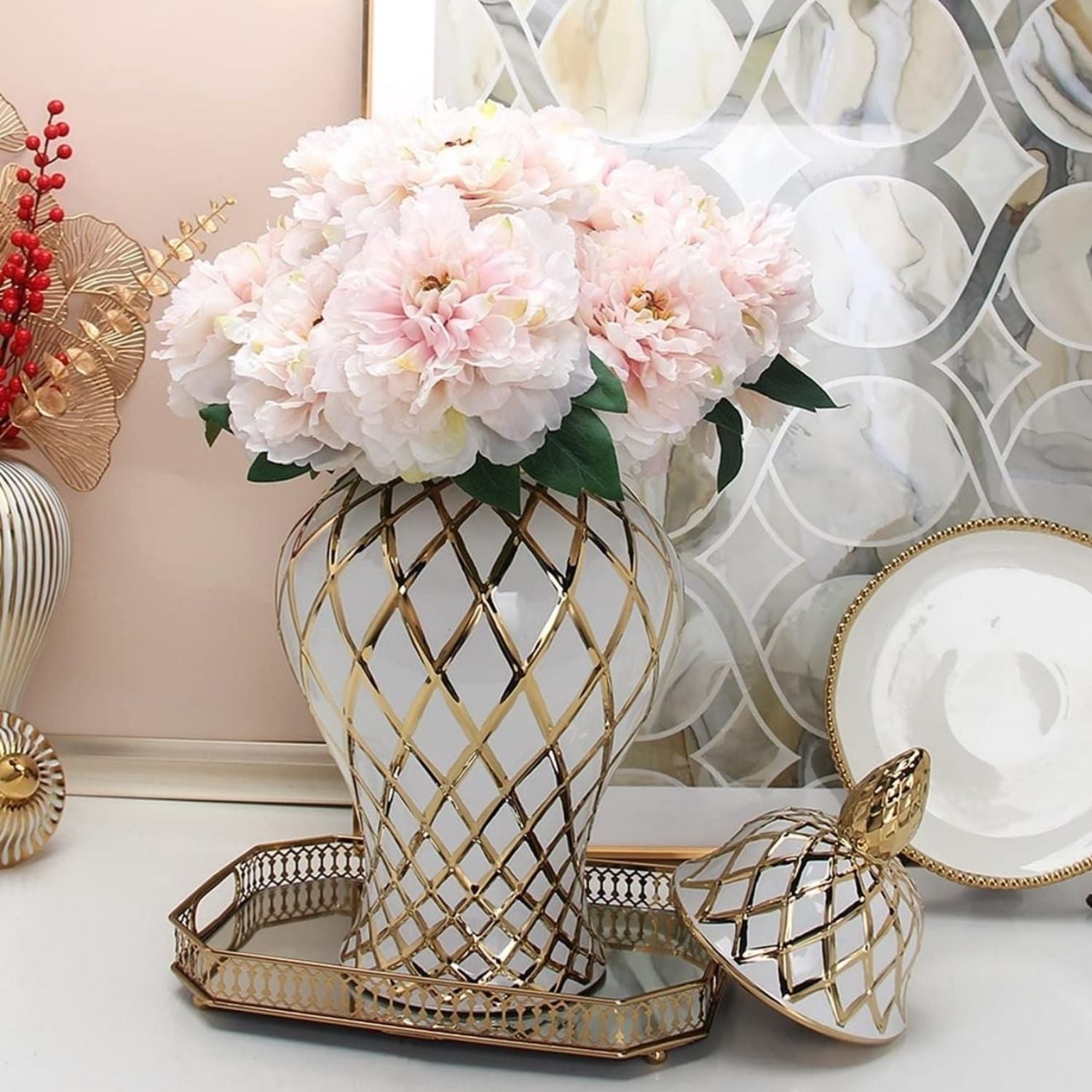 A decorative white ceramic vase with a gold lattice pattern, filled with pale pink artificial flowers and green leaves, displayed on a mirrored tray on a tabletop.