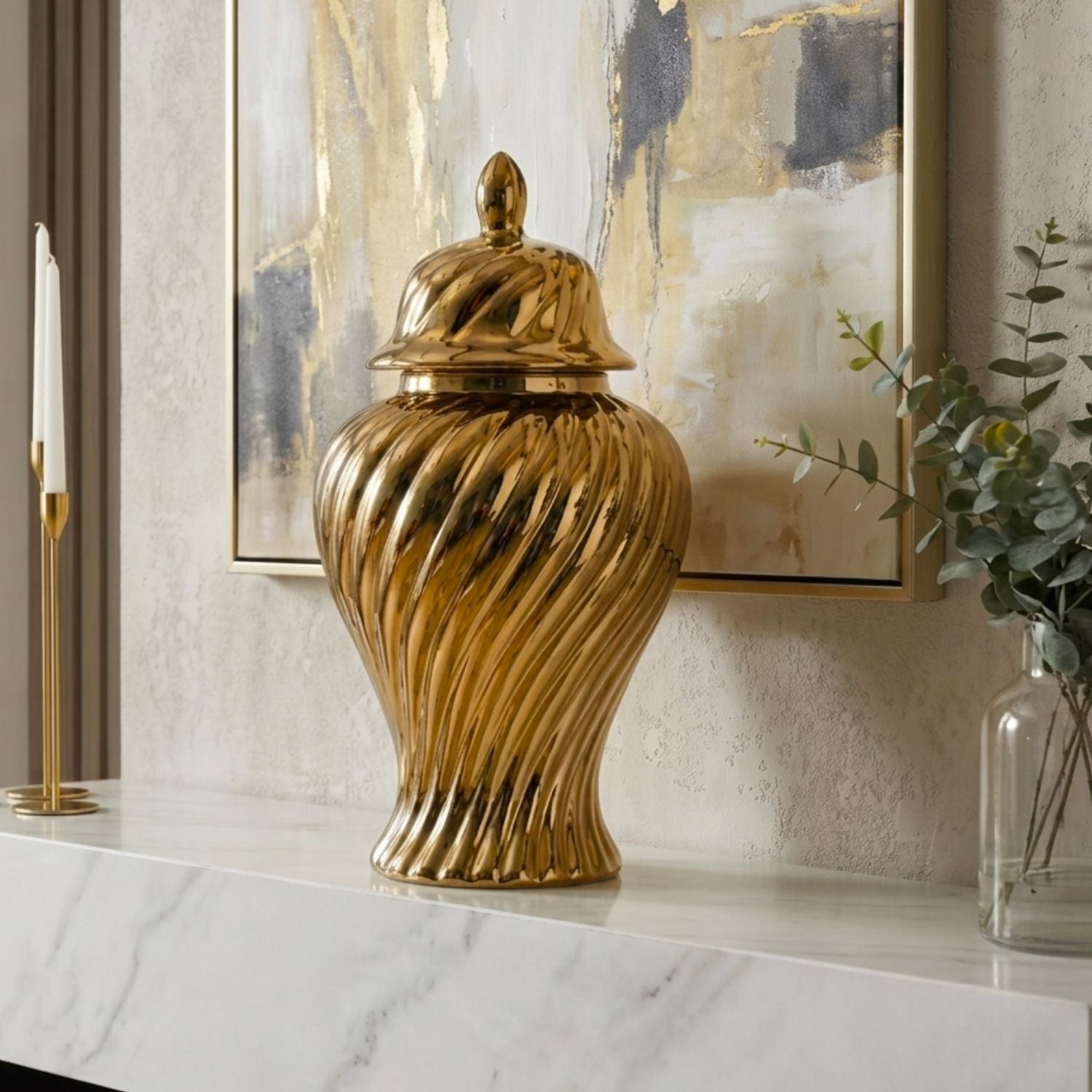 A large metallic gold decorative lidded jar with a ribbed, twisted surface sits on a white marble mantelpiece in a styled interior setting with a framed abstract painting, candles, and a glass vase.