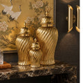 Three gold decorative ginger jars with ribbed swirl patterns and lids, arranged on a dark marble console table in an interior setting with a framed crane painting and wall sconces.