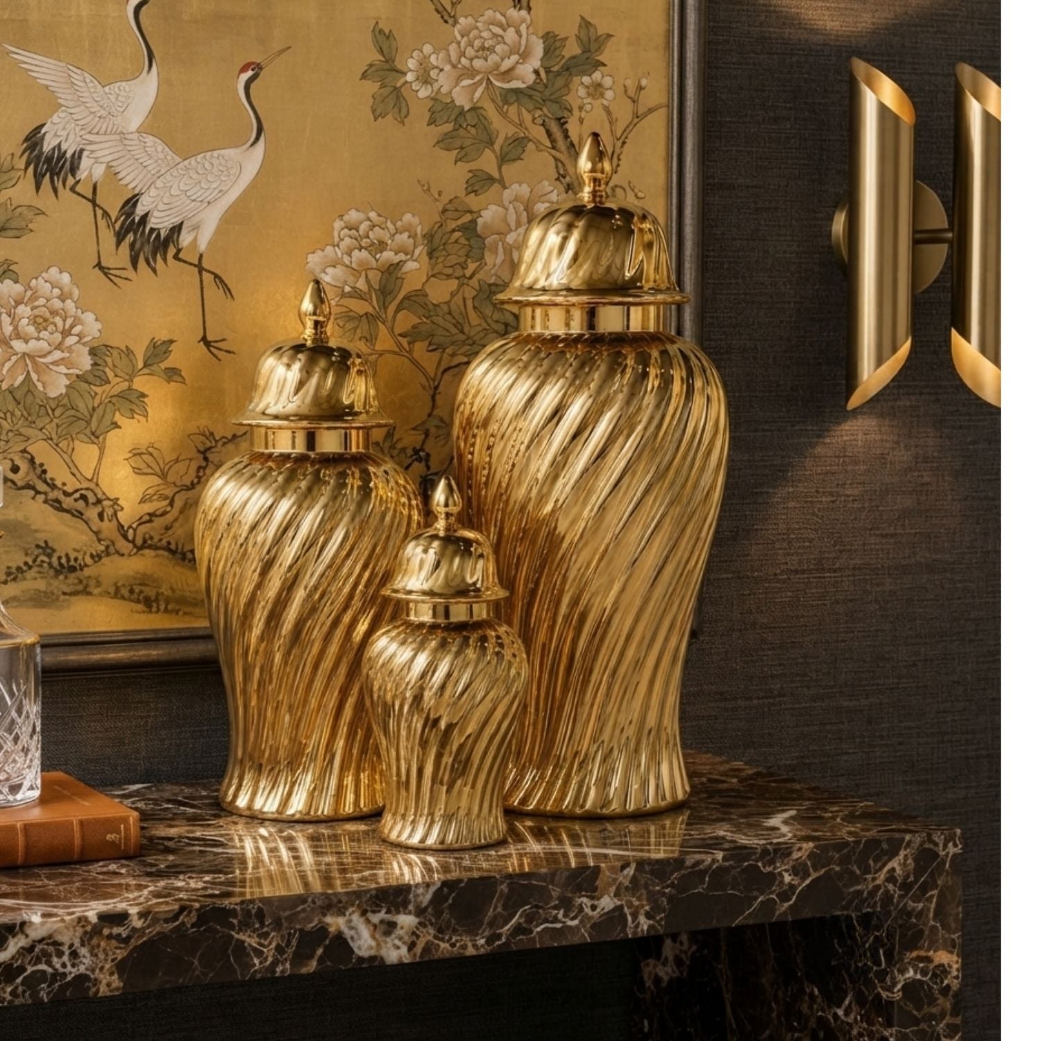 Three gold decorative ginger jars with ribbed swirl patterns and lids, arranged on a dark marble console table in an interior setting with a framed crane painting and wall sconces.