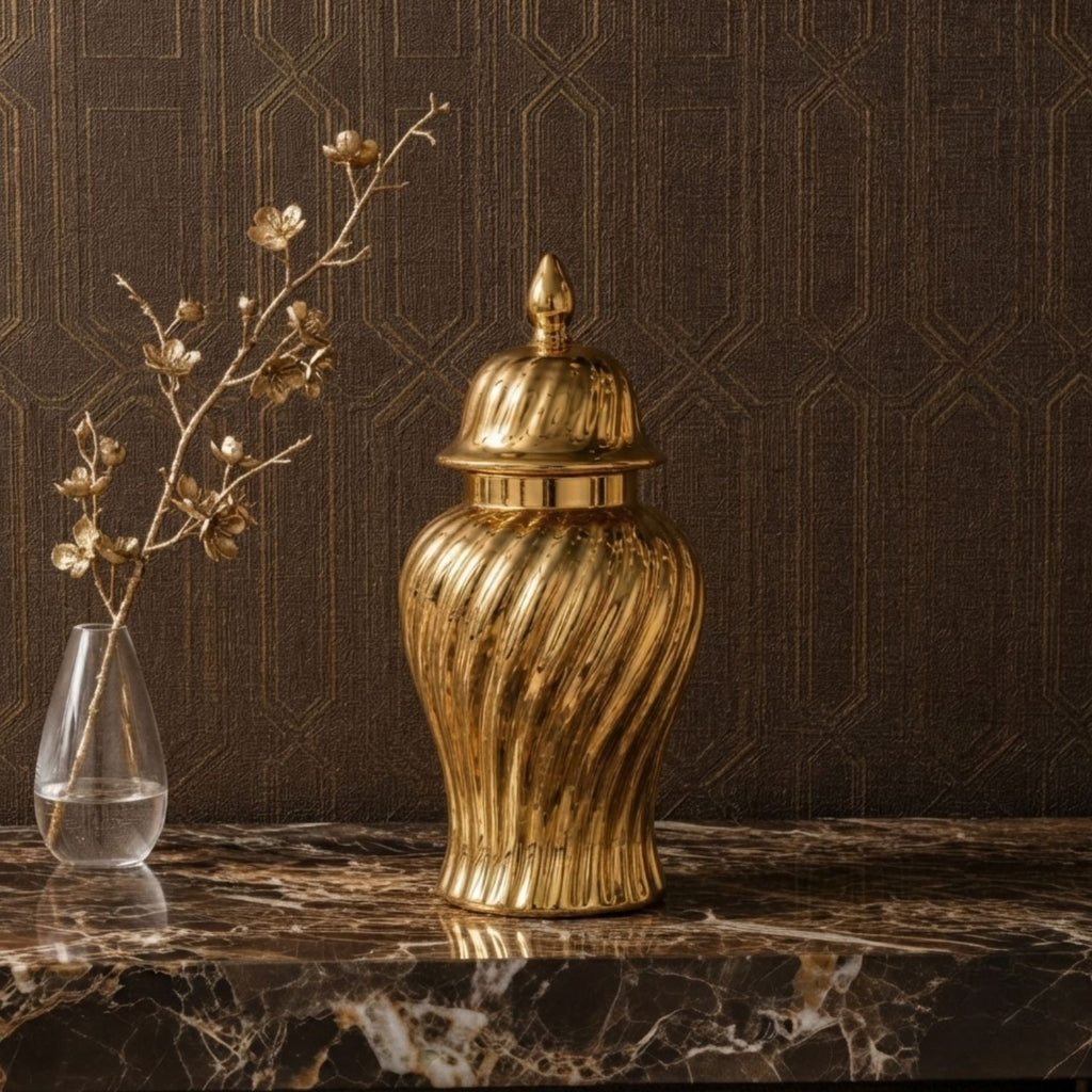 A metallic gold decorative jar with a ribbed, fluted body and a matching lid, displayed on a dark marble surface against a dark brown patterned wallpaper background.