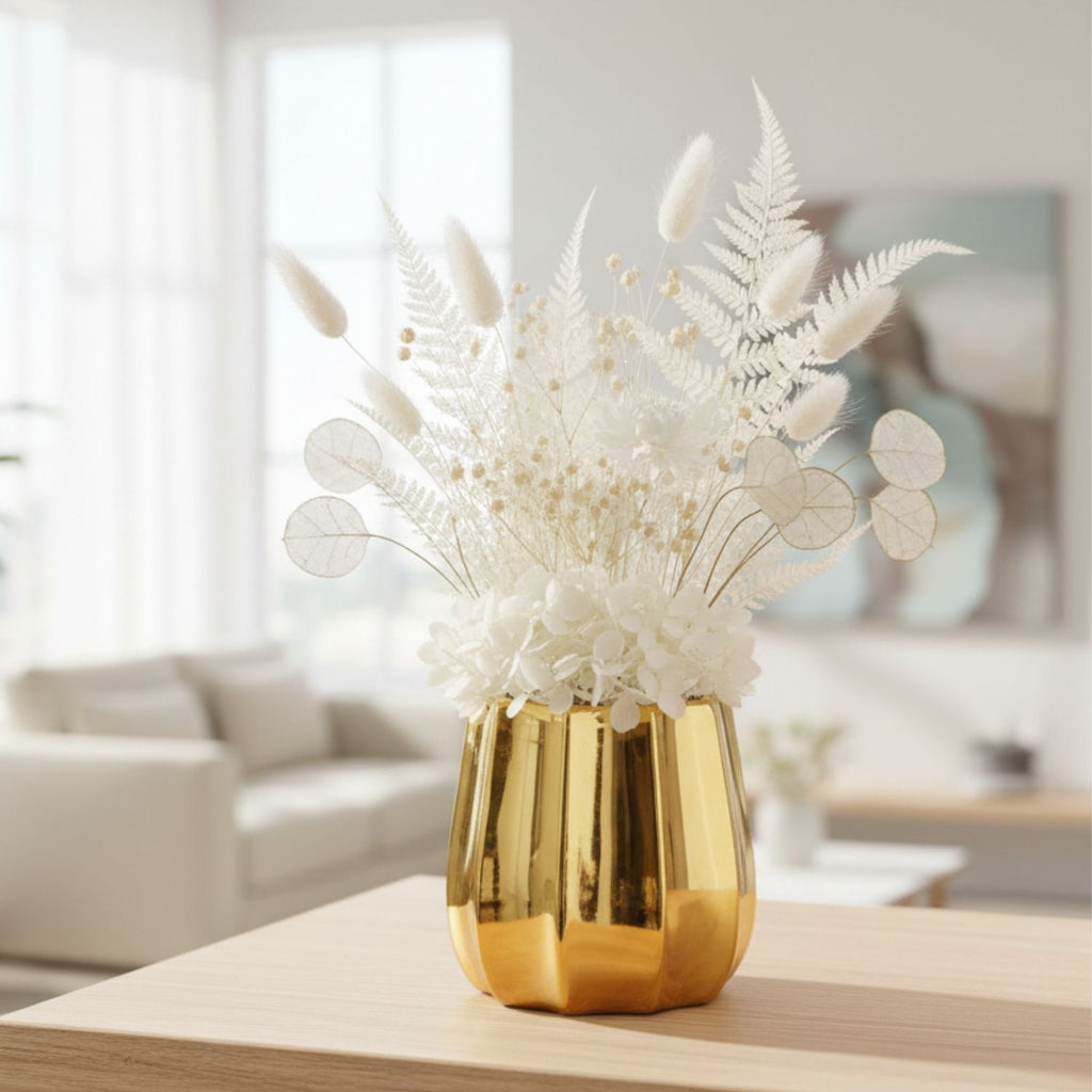 A shiny gold faceted vase filled with white dried flowers and ferns, displayed on a light wood table in a softly blurred living room setting.