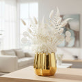 A shiny gold faceted vase filled with white dried flowers and ferns, displayed on a light wood table in a softly blurred living room setting.