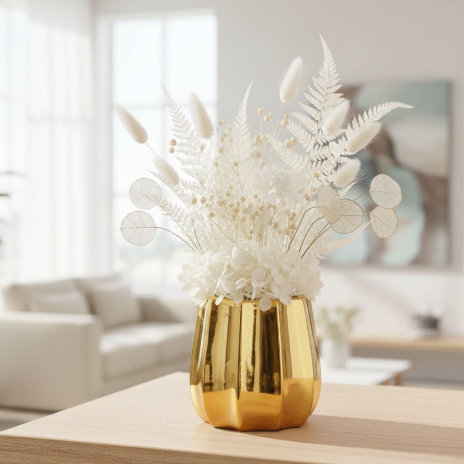 A shiny gold faceted vase filled with white dried flowers and ferns, displayed on a light wood table in a softly blurred living room setting.