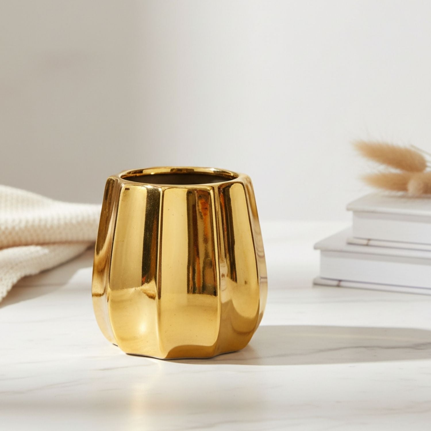 A shiny gold metallic planter with a faceted, ribbed design, sitting on a light marble surface with a neutral knit blanket and stacked books in the background.