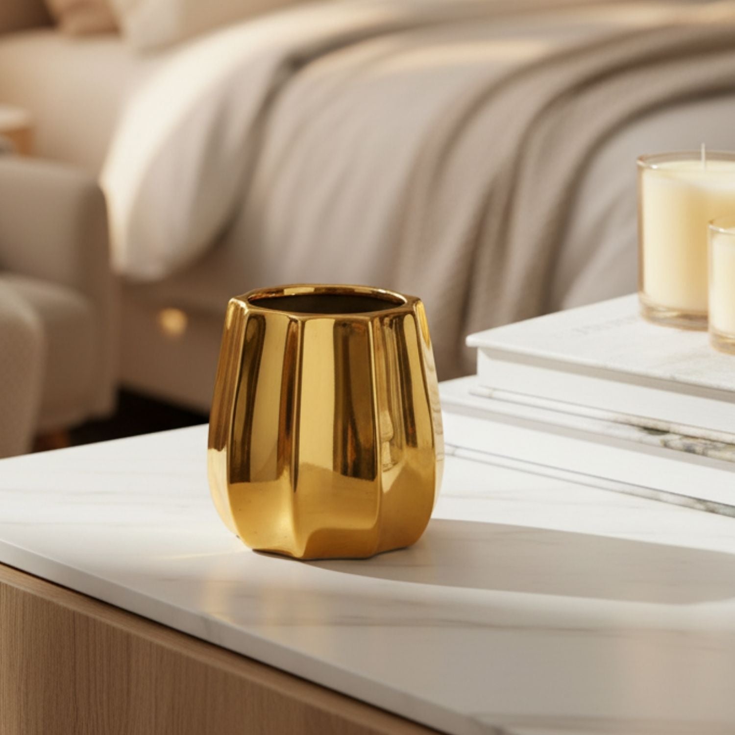 A shiny gold metallic cup or candle holder with a faceted, ribbed design sits on a white marble table in a softly lit bedroom setting.