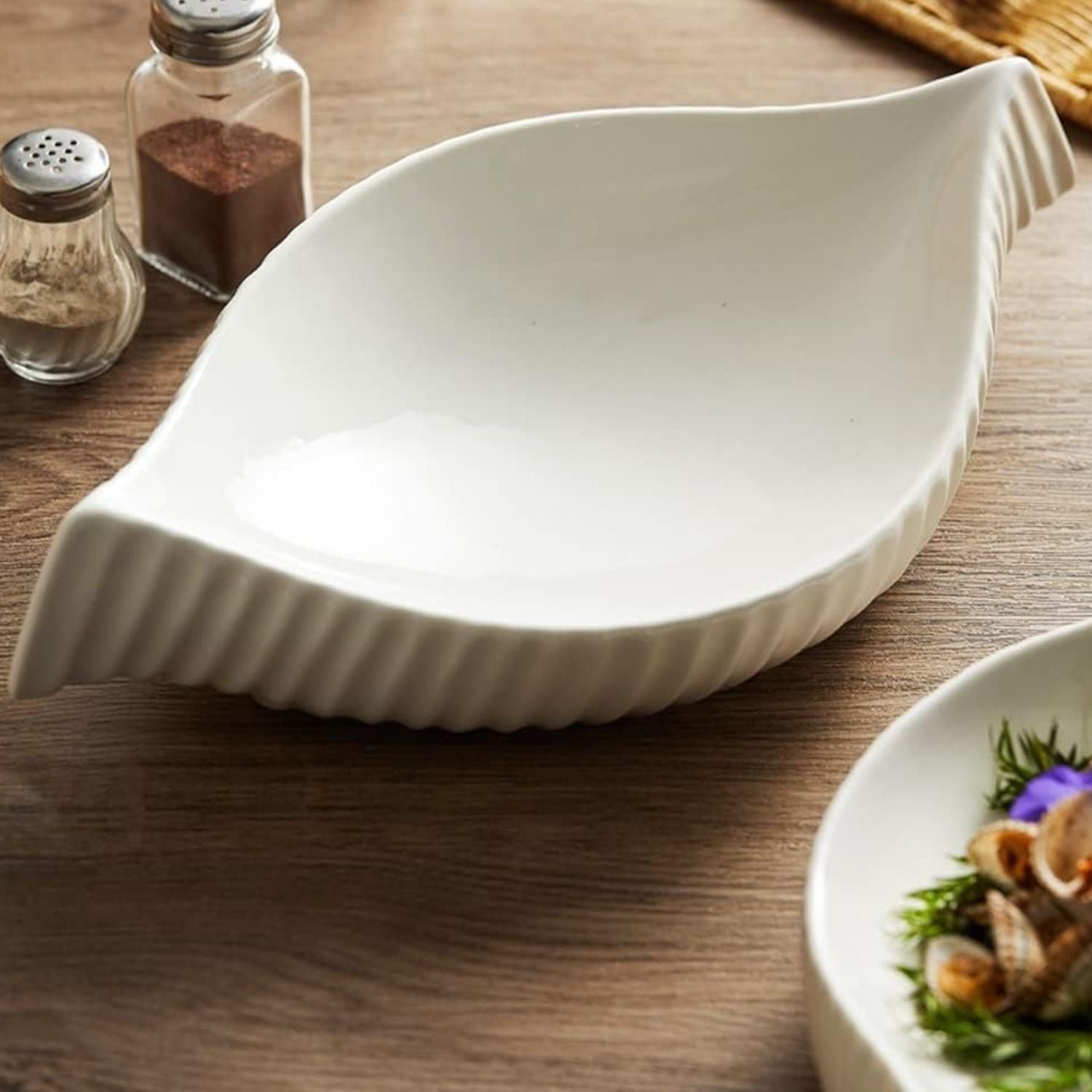 A white ceramic leaf-shaped serving bowl with ribbed flared edges, shown on a wooden tabletop with salt and pepper shakers and part of a plated dish nearby.