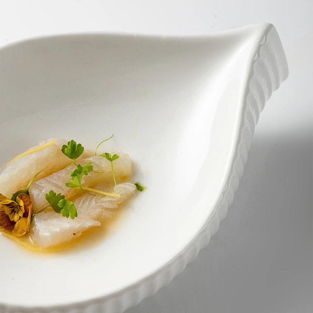 A close-up of a white ceramic dish with a leaf-like shape and scalloped rim, holding a small serving of pale fish or seafood in a light broth with green herb garnish on a white background.