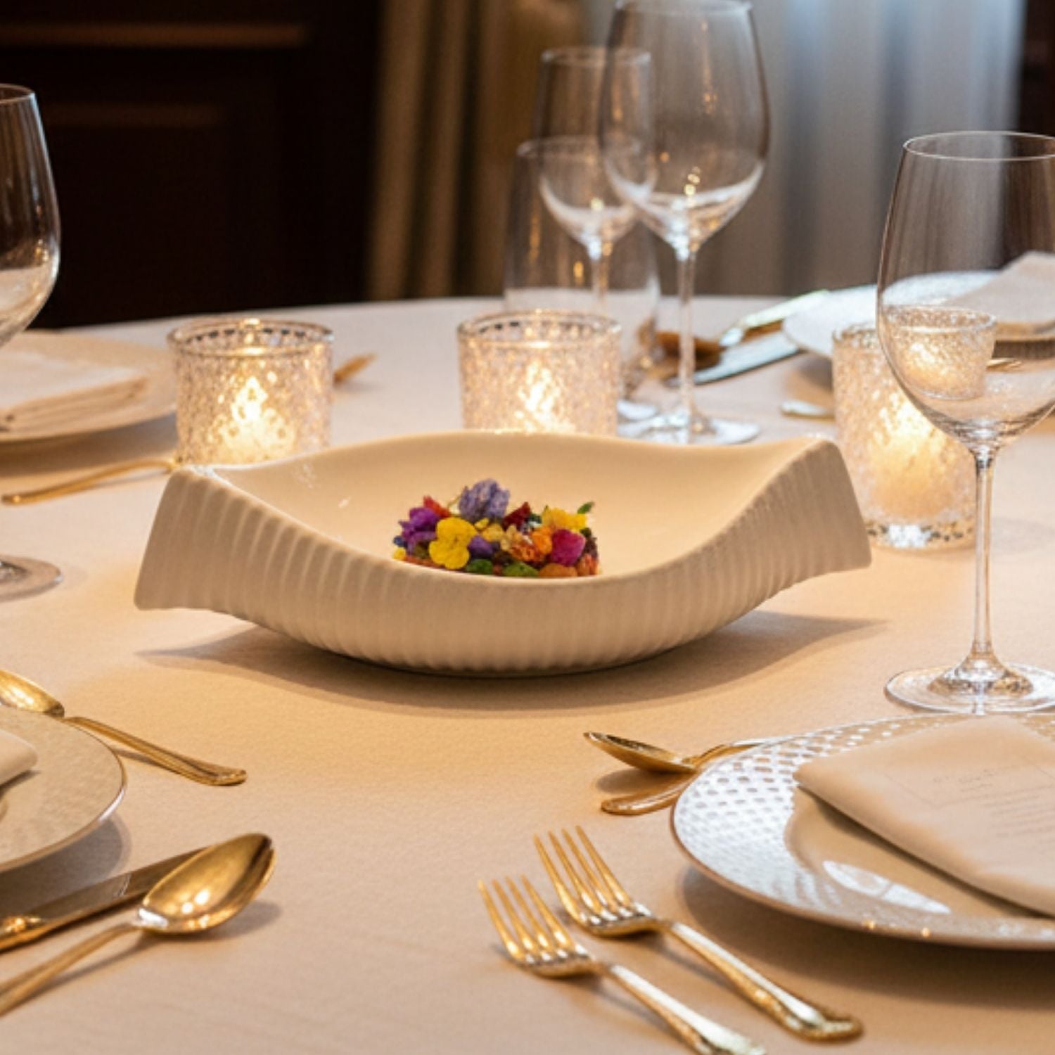 A white ribbed ceramic bowl with a wavy rim, styled on a beige tablecloth with gold cutlery, wine glasses, and lit candle holders.