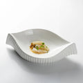 A white ceramic leaf-shaped serving dish with ribbed flared edges, shown on a clean white background with a small plated food garnish inside.