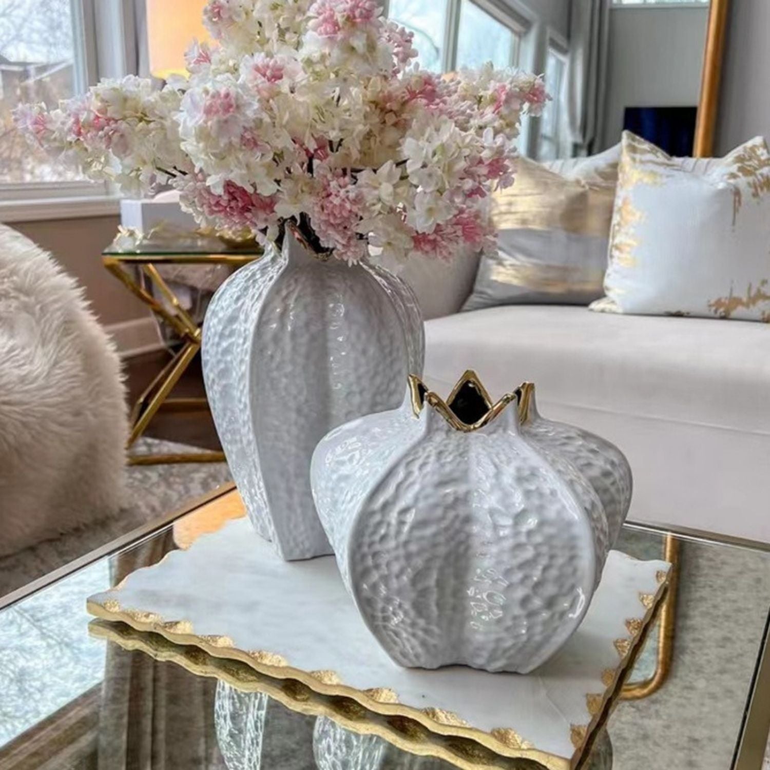 Two textured ceramic pumpkin-shaped vases in white, one tall and one short, displayed on a glass tray table in a bright living room setting with pink and white flowers.