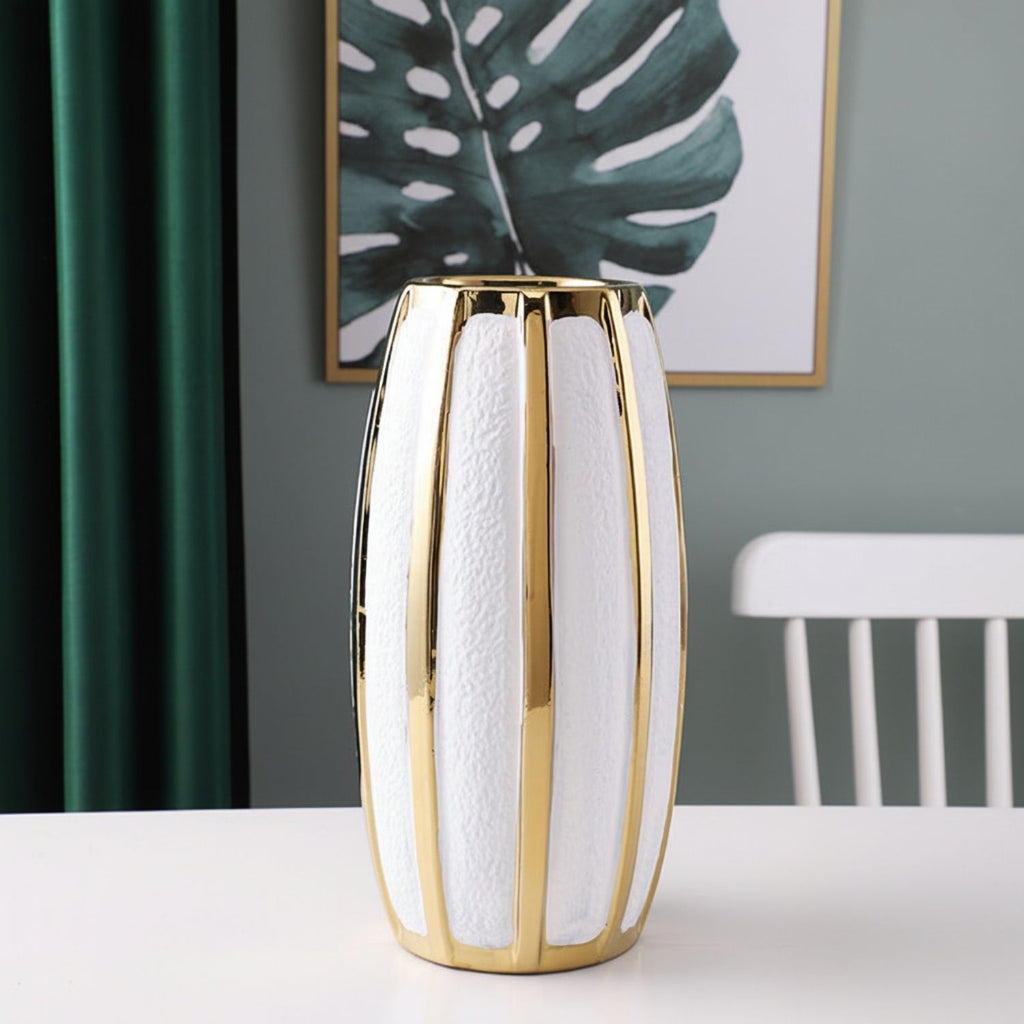 A modern cylindrical vase with a textured white body and polished gold vertical stripes, displayed on a white table in a styled room setting with a green curtain and framed wall art in the background.