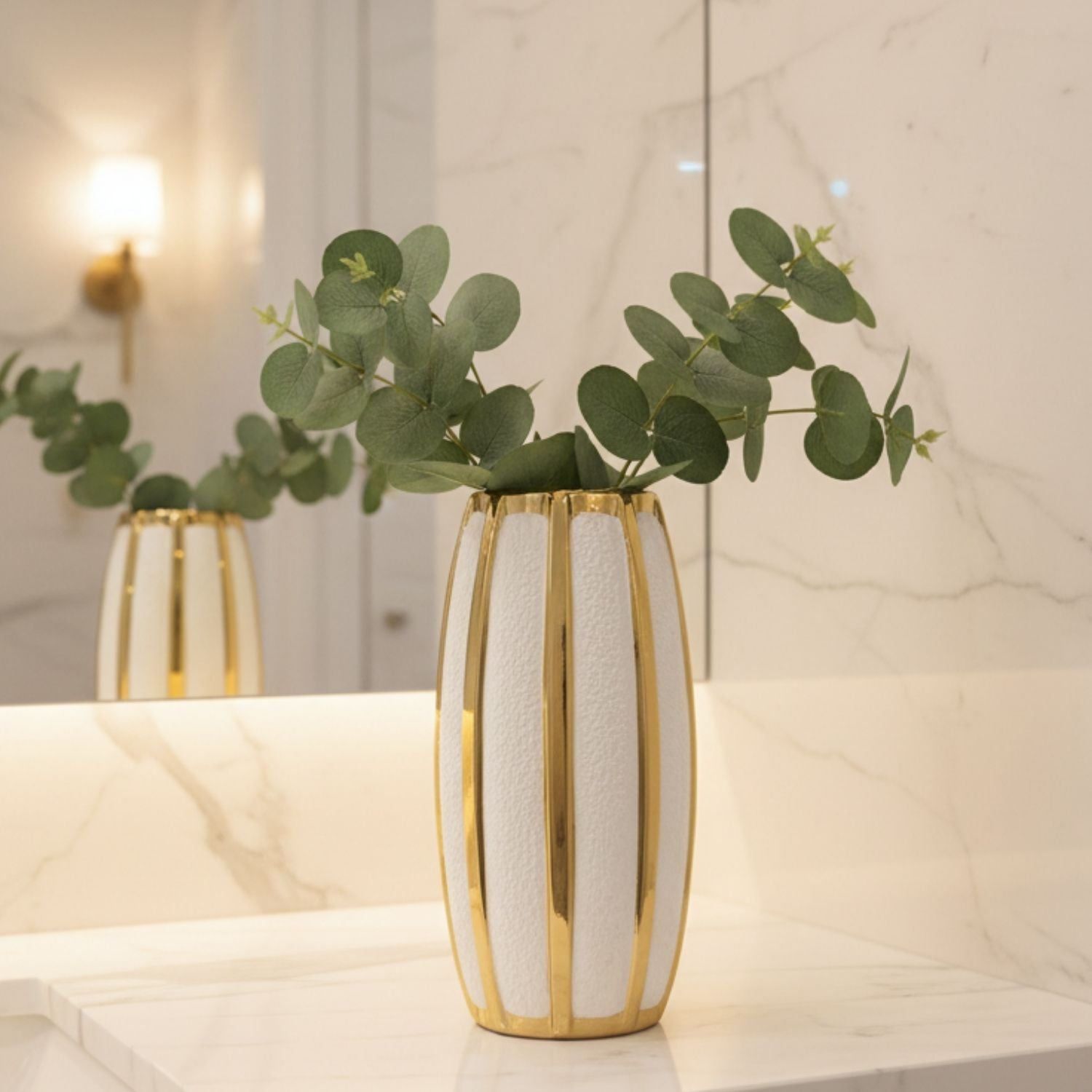 A modern cylindrical vase with vertical gold metallic stripes and a textured white center, filled with green eucalyptus branches, set on a white marble countertop with a matching marble backsplash and a mirror in the background.