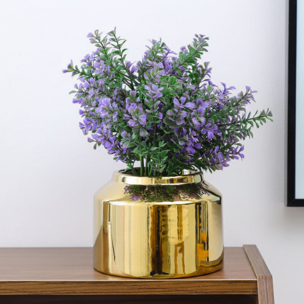 A shiny gold cylindrical vase filled with artificial purple flowers and green leaves, displayed on a wooden surface against a plain light wall.