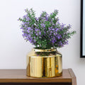A shiny gold cylindrical vase filled with artificial purple flowers and green leaves, displayed on a wooden surface against a plain light wall.