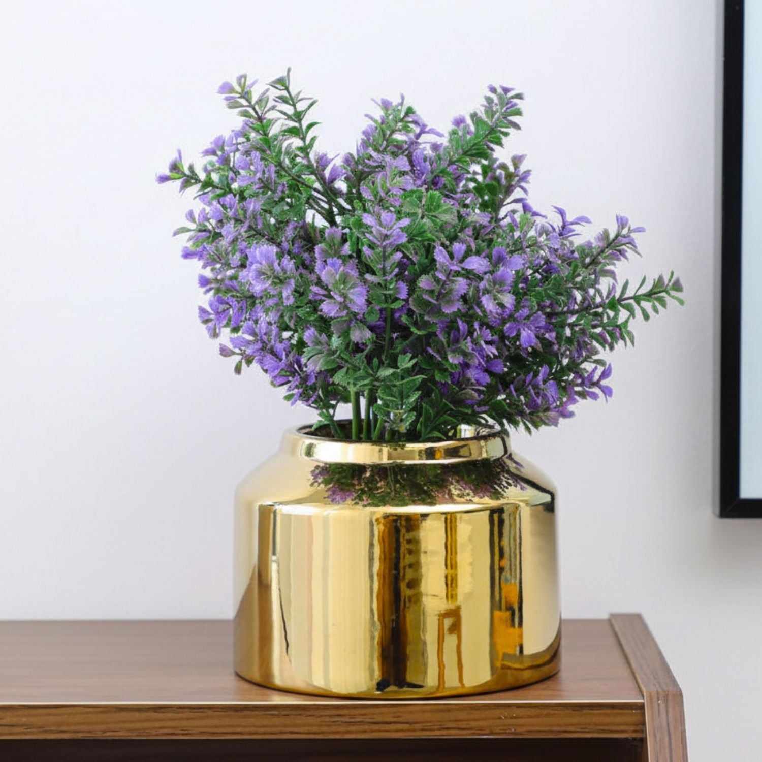 A shiny gold cylindrical vase filled with artificial purple flowers and green leaves, displayed on a wooden surface against a plain light wall.