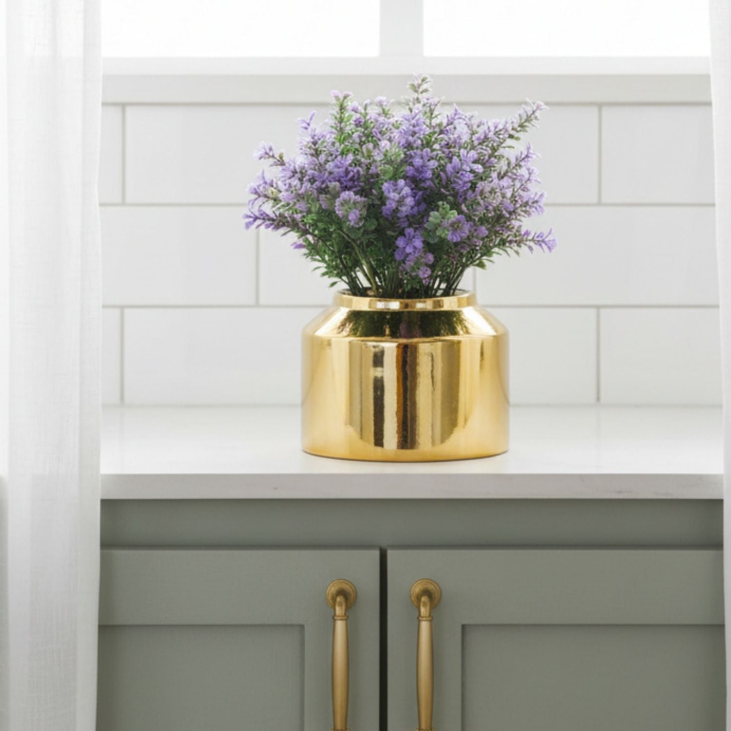 A shiny gold cylindrical vase filled with purple flowers and green leaves, displayed on a white countertop in front of a white subway tile backsplash.